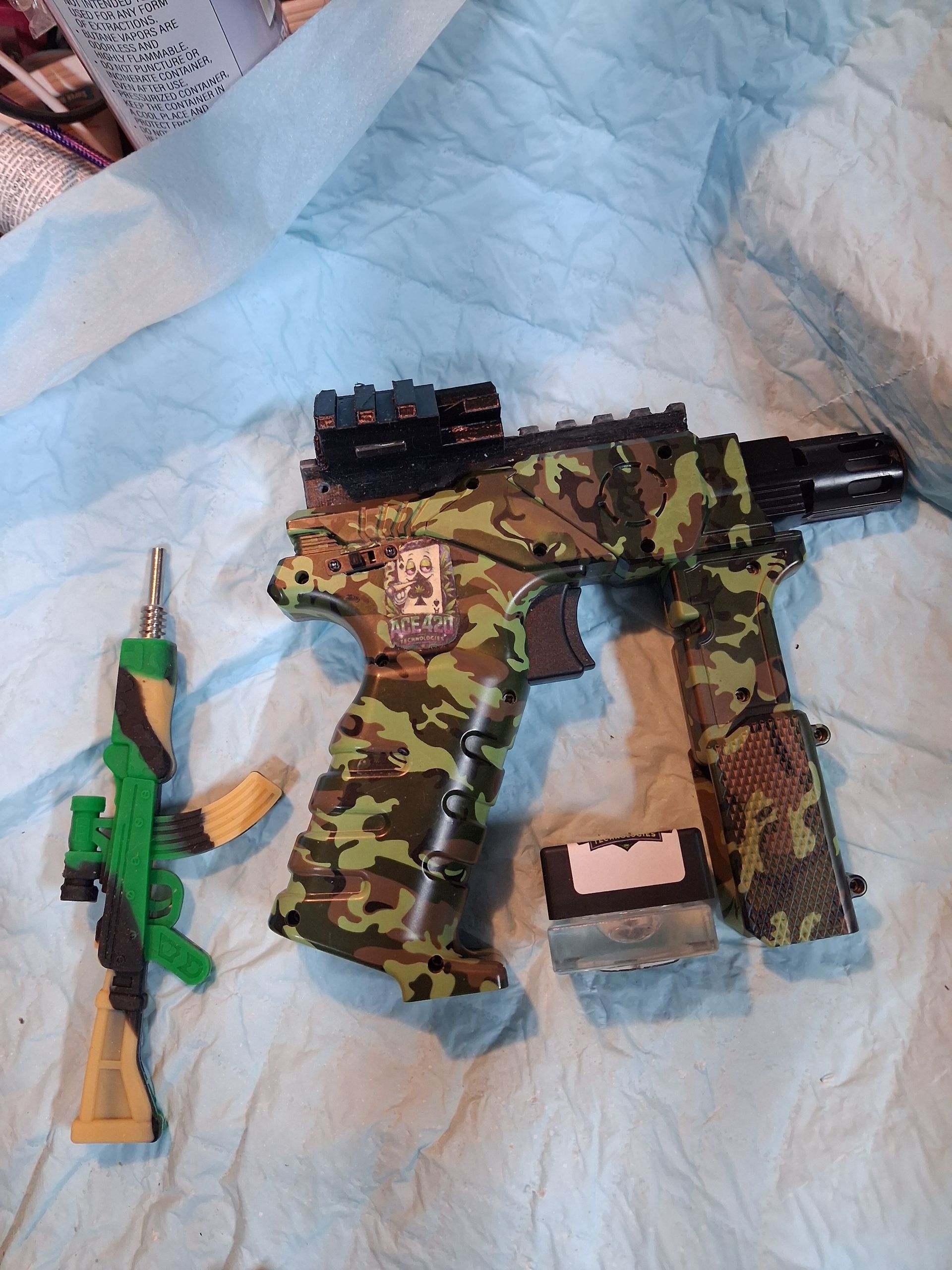 Camouflage toy gun with green-and-orange toy beside it on a white blanket.