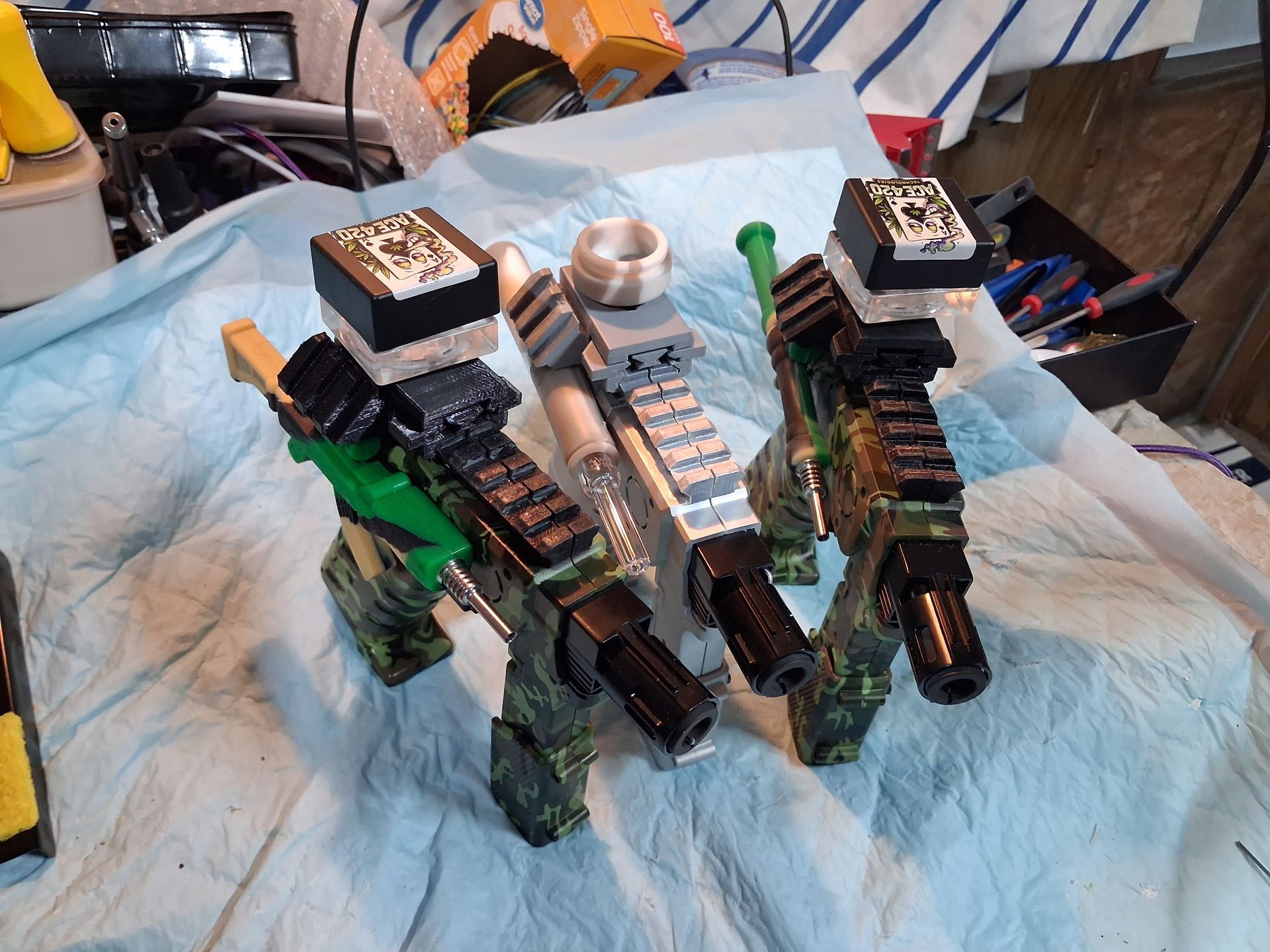 3 blaster shaped Butane Torches in silver and camouflage  with green, black, and white parts on a blue cloth-covered table