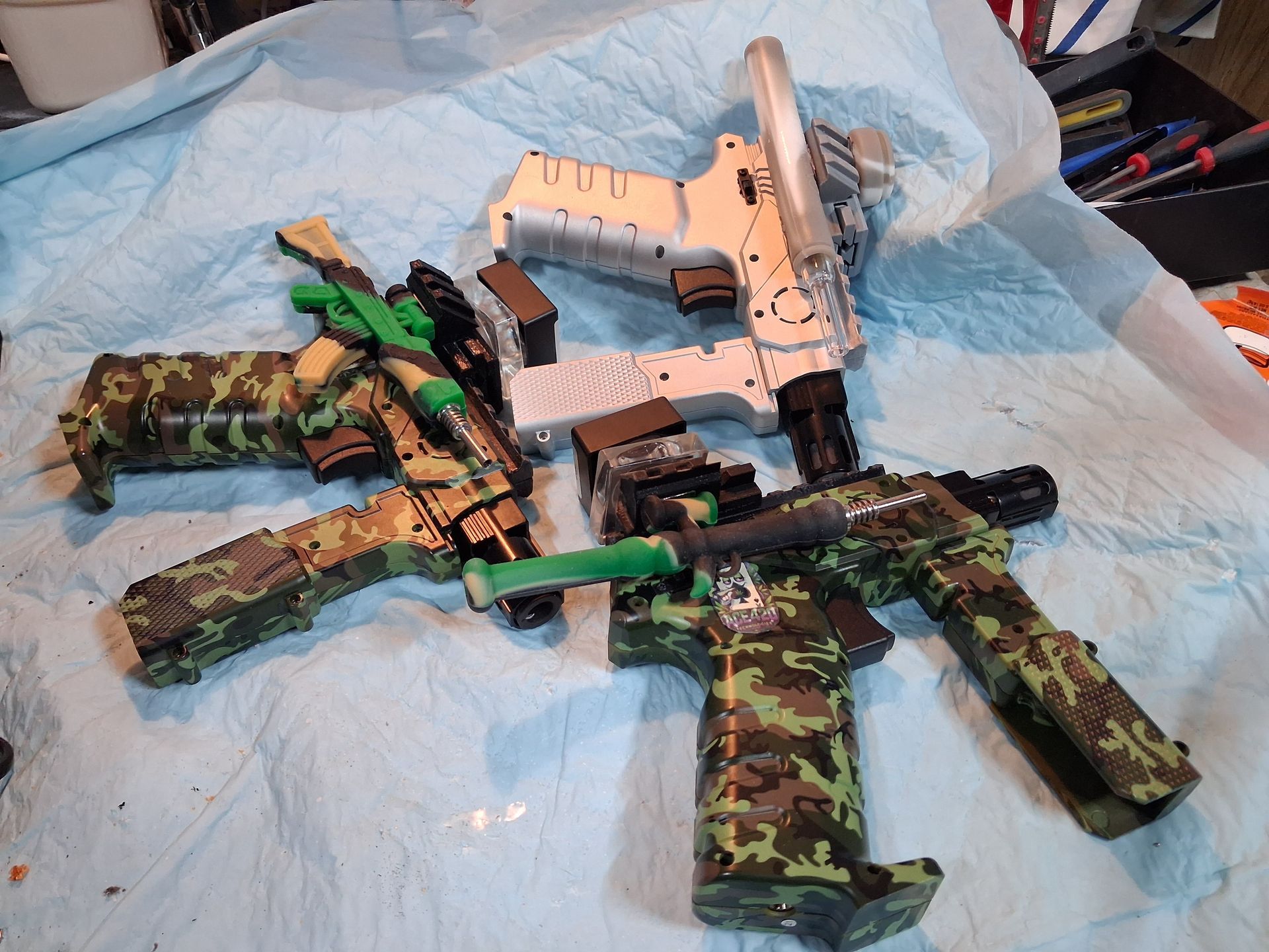 Camouflage-patterned toy gun shaped torches arranged on a blue sheet, including one silver torch and two green rifles