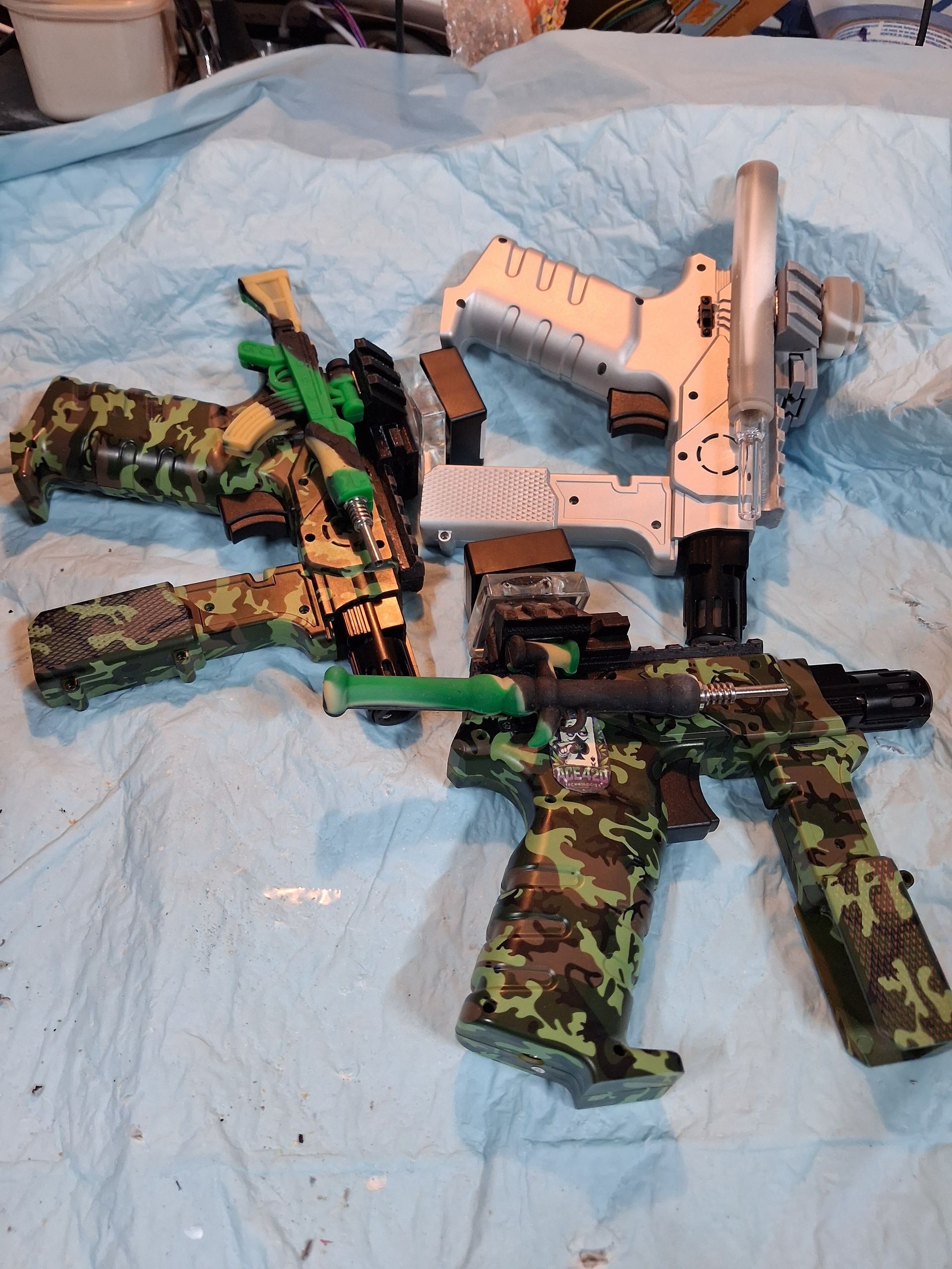 Camouflaged toy guns and a white toy pistol laid on a blue cloth background