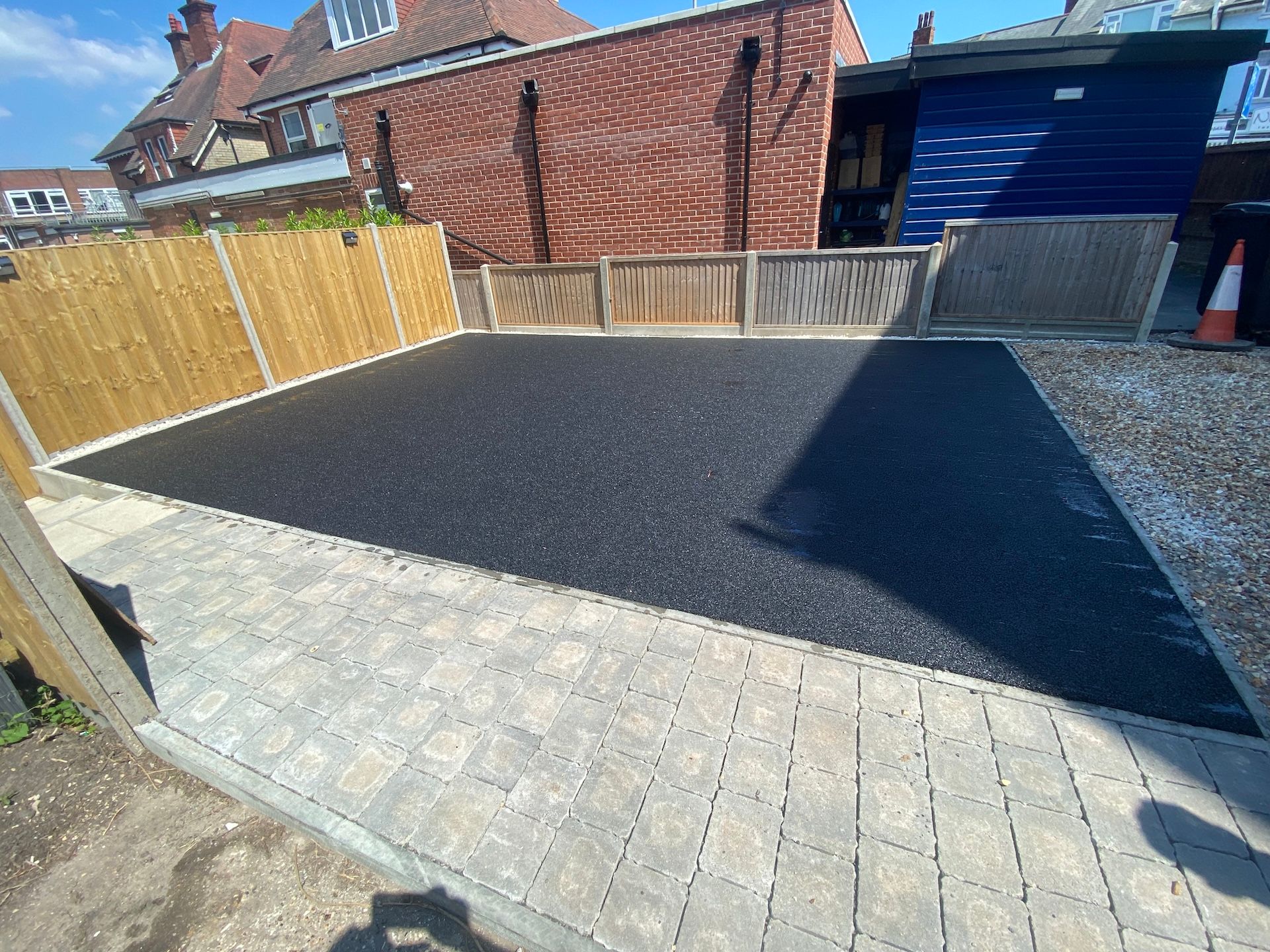 A black asphalt driveway with a wooden fence and a brick building in the background.