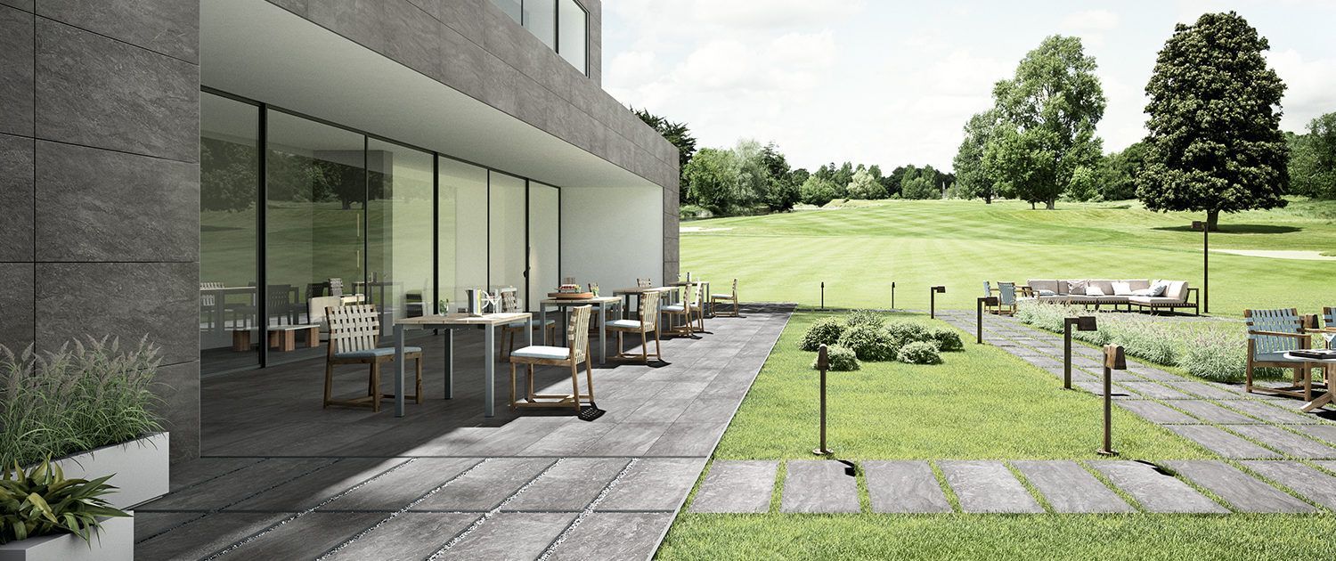 Eine Terrasse mit Natursteinen, Tischen und Stühlen vor einem Gebäude mit einem Golfplatz im Hintergrund.
