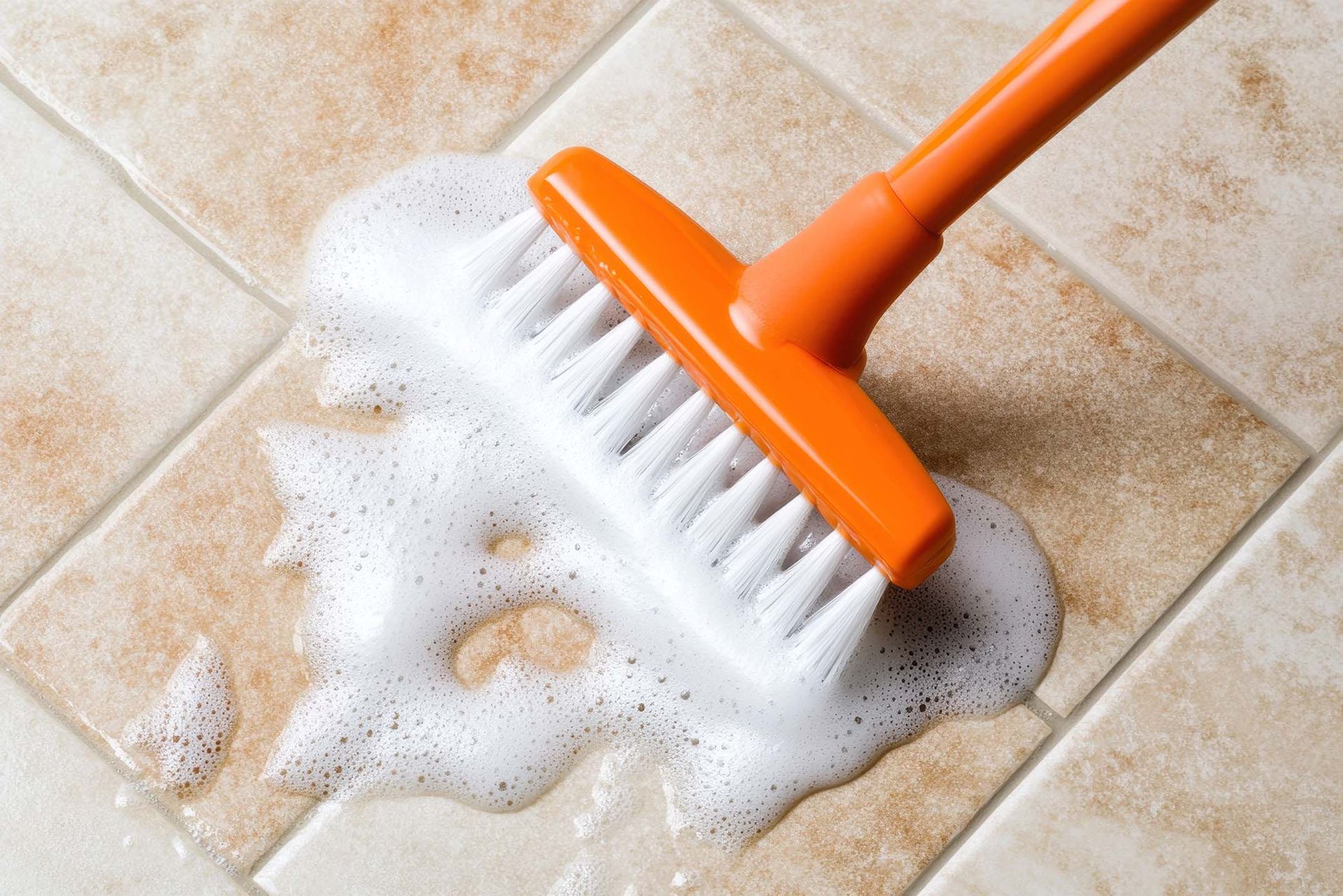 Eine orangefarbene Bürste reinigt einen Fliesenboden mit Seife
