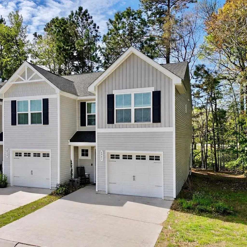Two-story gray townhome with two garages, white trim, and dark shutters, set against a wooded backdrop under a blue sky.