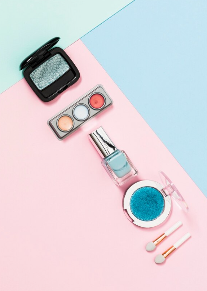 Cosméticos dispuestos sobre un fondo rosa, azul y verde: sombras de ojos, esmaltes de uñas, brochas y estuches de maquillaje.