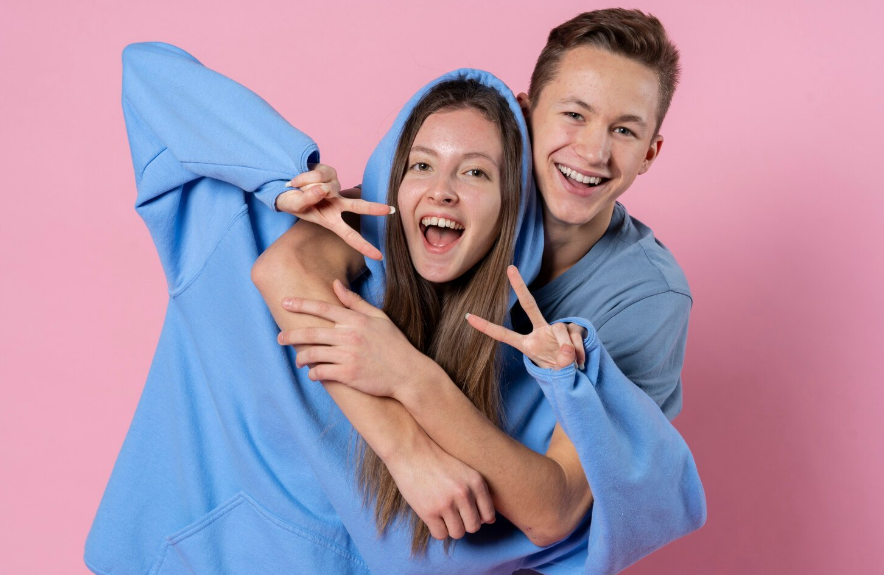 Dos personas abrazadas, con sudaderas azules, haciendo el signo de la paz y sonriendo sobre un fondo rosa.