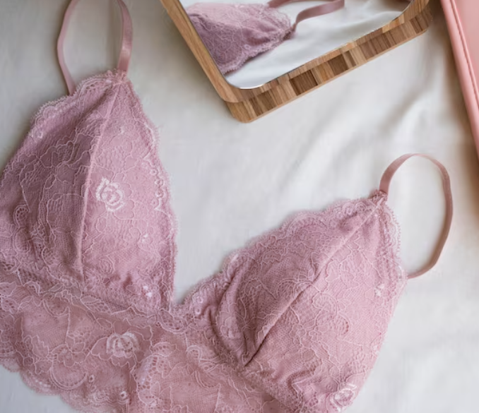 Sujetador de encaje rosa sobre tela blanca, con un espejo y un objeto rosa al fondo.