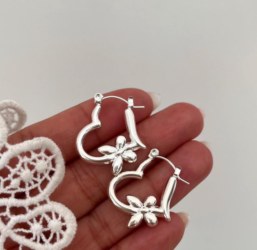 Pendientes de aro plateados en forma de corazón, cada uno con un dije de flor, sostenidos en la mano.