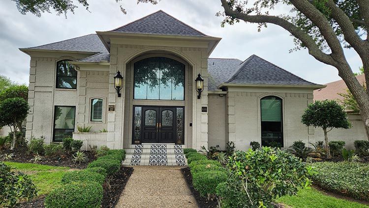 Beige brick home with dark roof, arched entryway, and landscaped front yard.