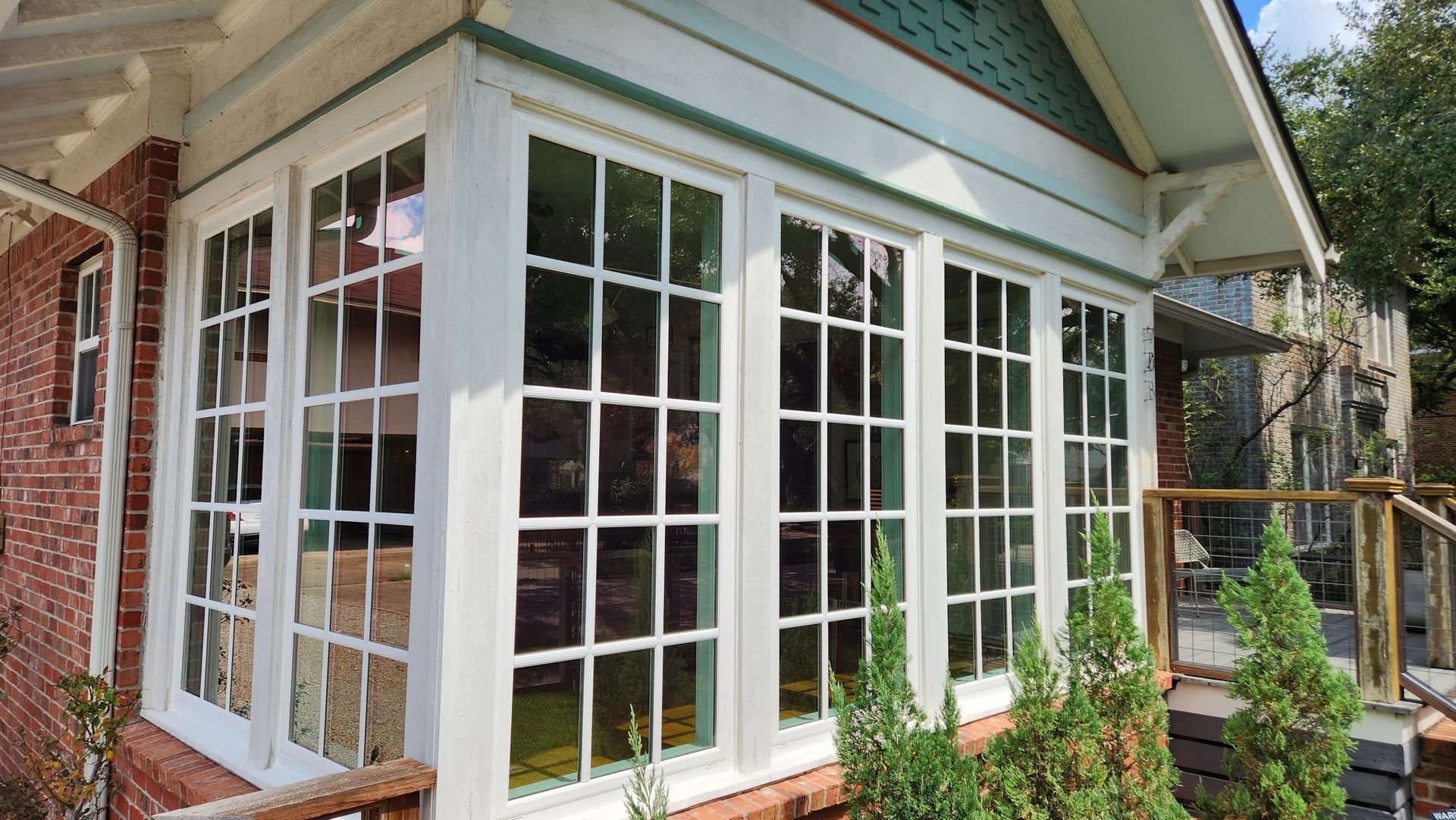 White-framed windows of a sunroom on a brick building, green trim, small evergreen plants in front.