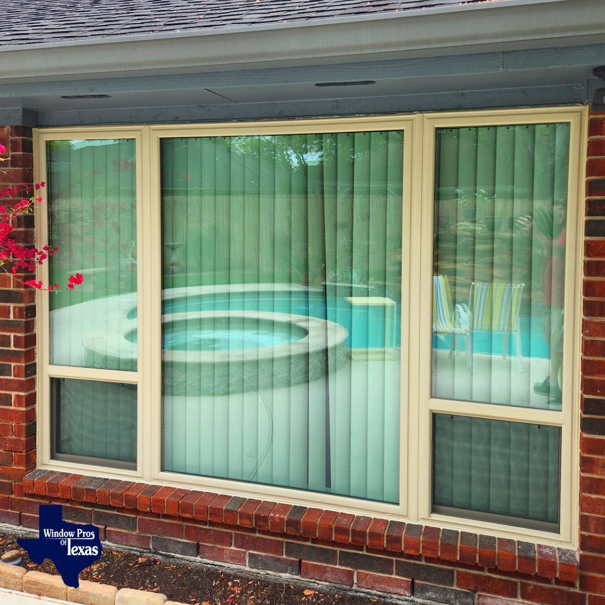 Exterior view of a window with vertical blinds, overlooking a pool. Brick wall with a beige window frame.