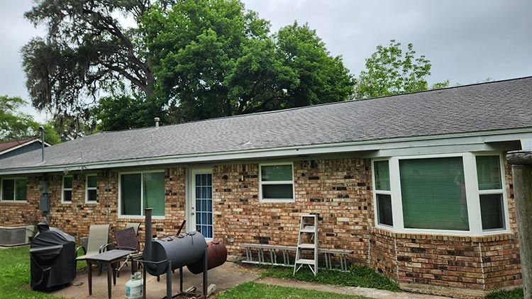 Brick house with dark roof and green trees; backyard with patio and barbecue grills.