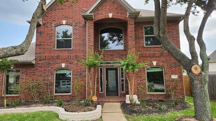Two-story brick house with dark door, arched entryway, and large windows, surrounded by trees and landscaping.