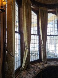 Alt text: A photo of three golden and cream curtains, tied back in the middle to show the leaded art glass windows. A closeup of the larkspur pattern on the curtain.
