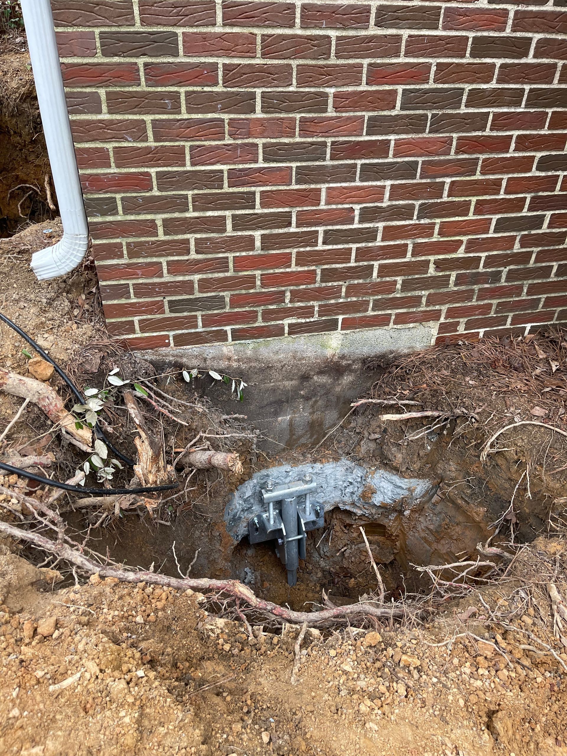 A foundation with exposed plumbing in a dug-out area next to a red brick wall. A white downspout is visible.