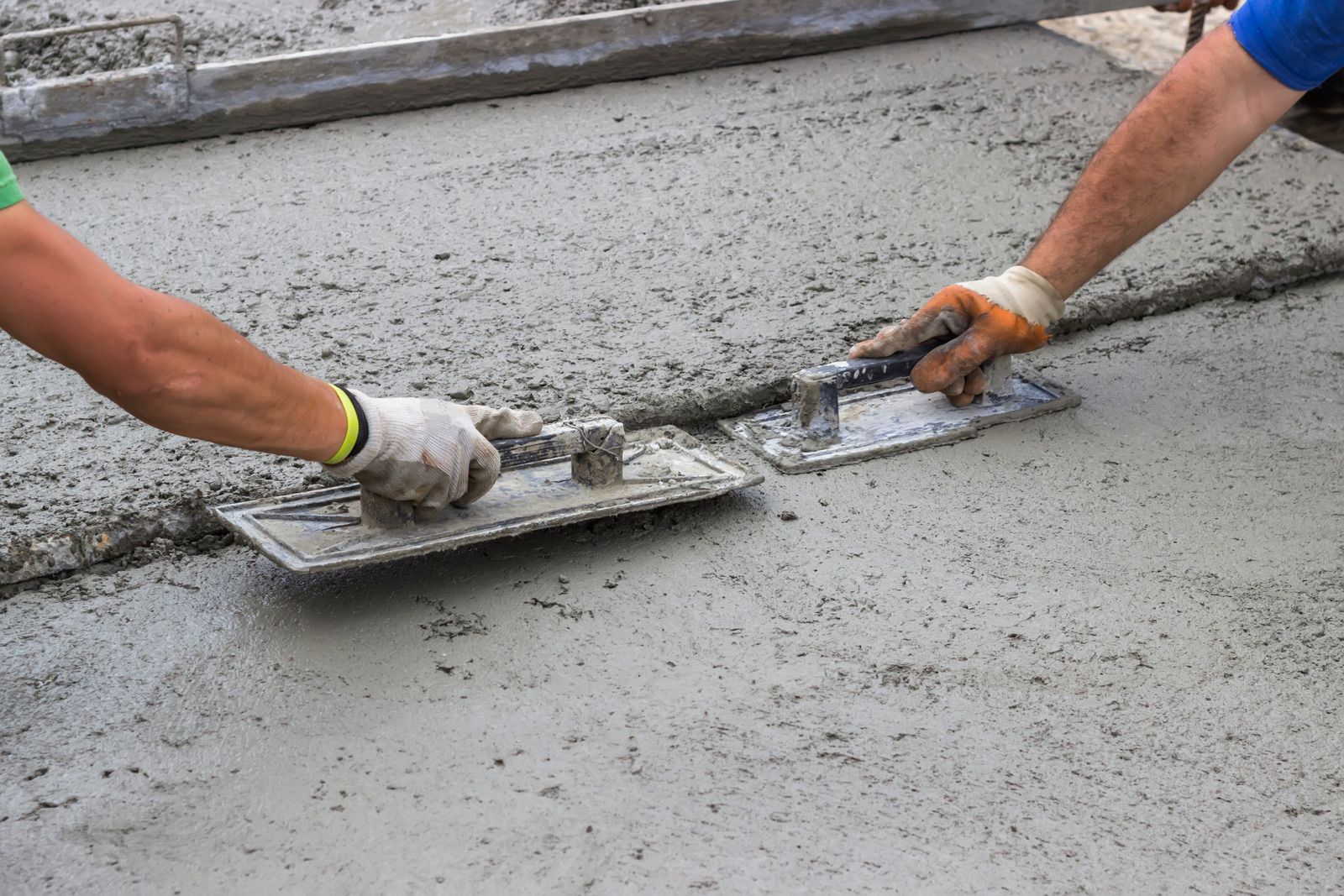 Two workers smoothing wet concrete with trowels outdoors.