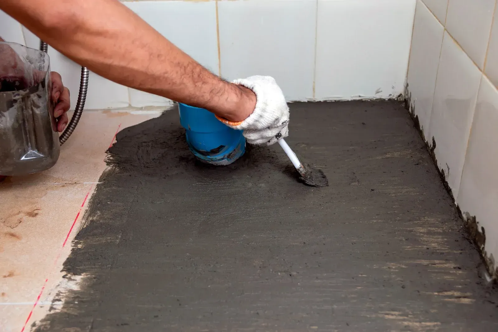 Person in a glove troweling gray cement in a bathroom, near a blue bucket.