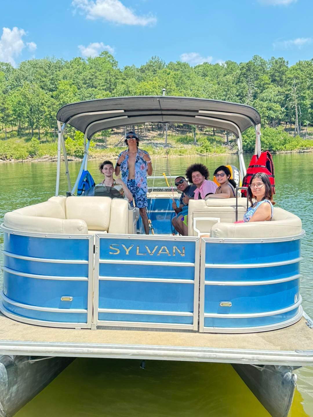 A group of people sitting on a blue Sylvan pontoon boat on a sunny lake with trees in the background.