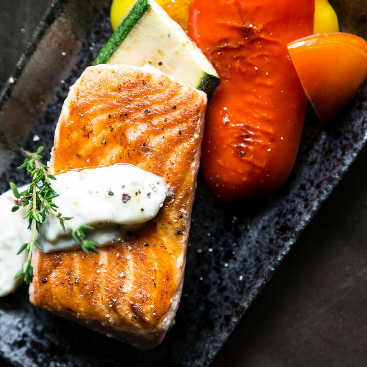 香煎鮭魚佐白醬，烤蔬菜，盛於深色盤子中。