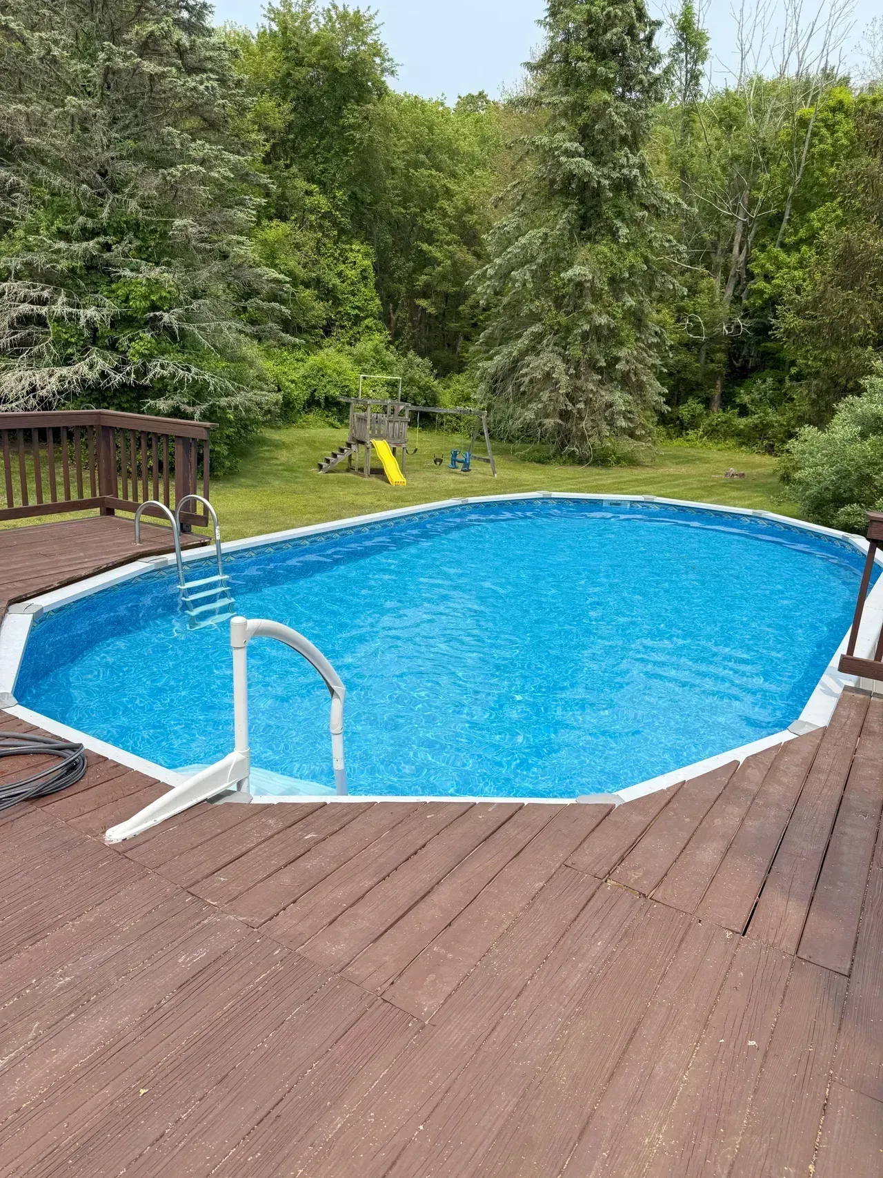 Backyard swimming pool with blue water, wooden deck, and surrounding trees