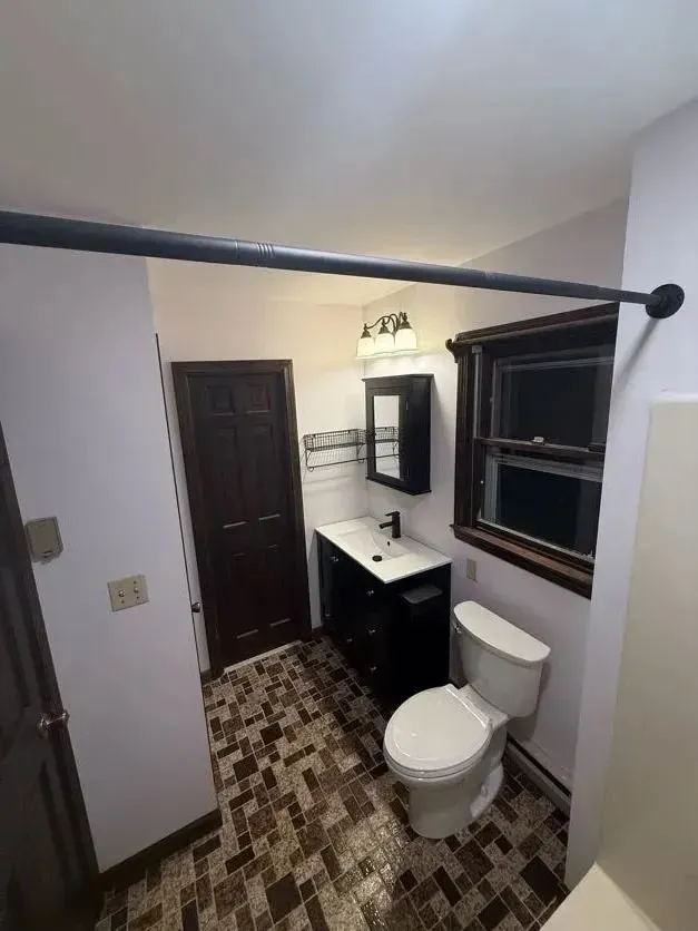 Small bathroom with toilet, vanity sink, mirror, and window in a narrow tiled room.