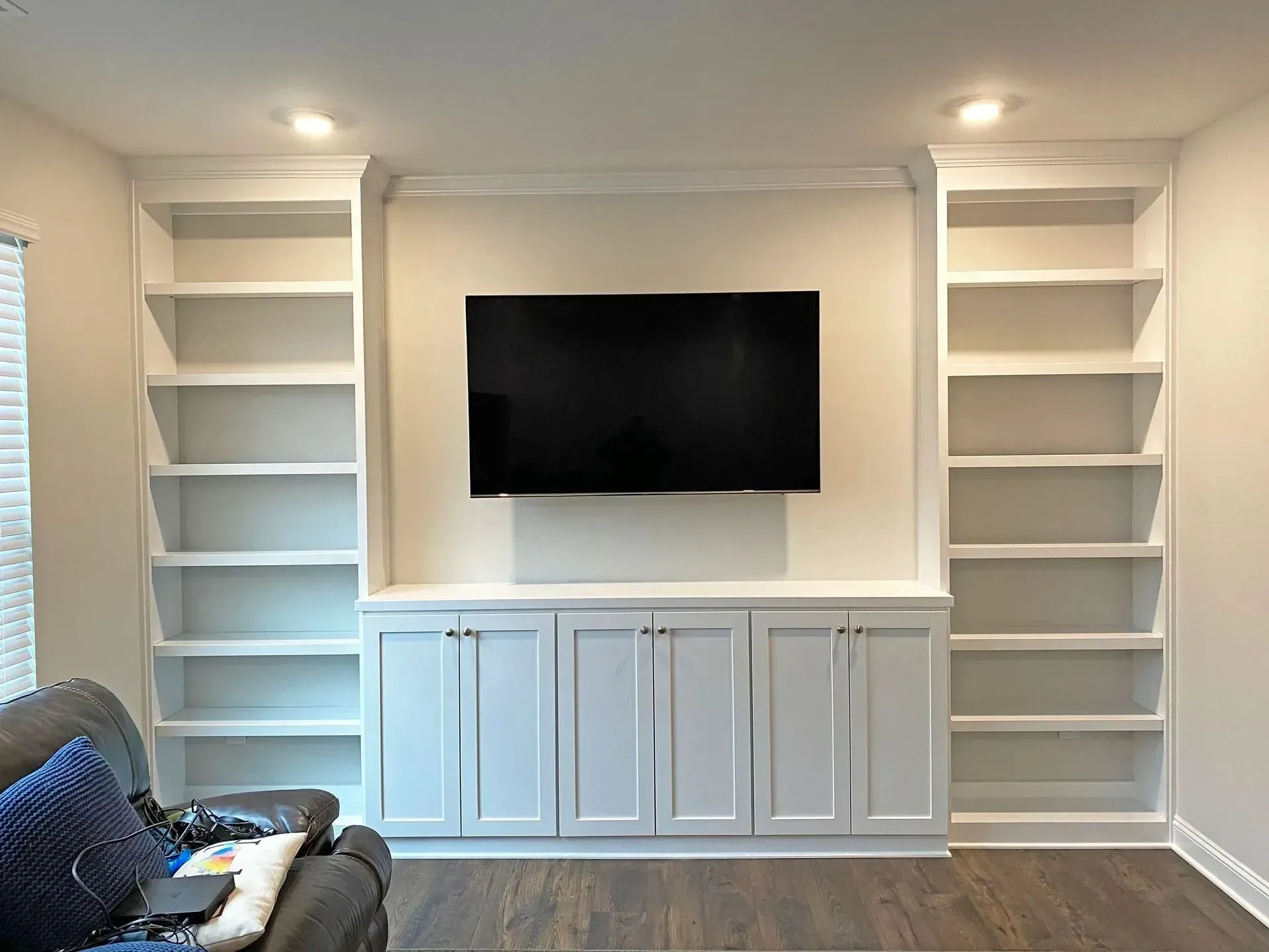 Modern living room wall with mounted TV, white cabinets, built-in shelves, and a chair on the left.