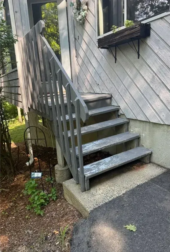 Outdoor wooden stairs with railing beside a house, leading up from a paved path.
