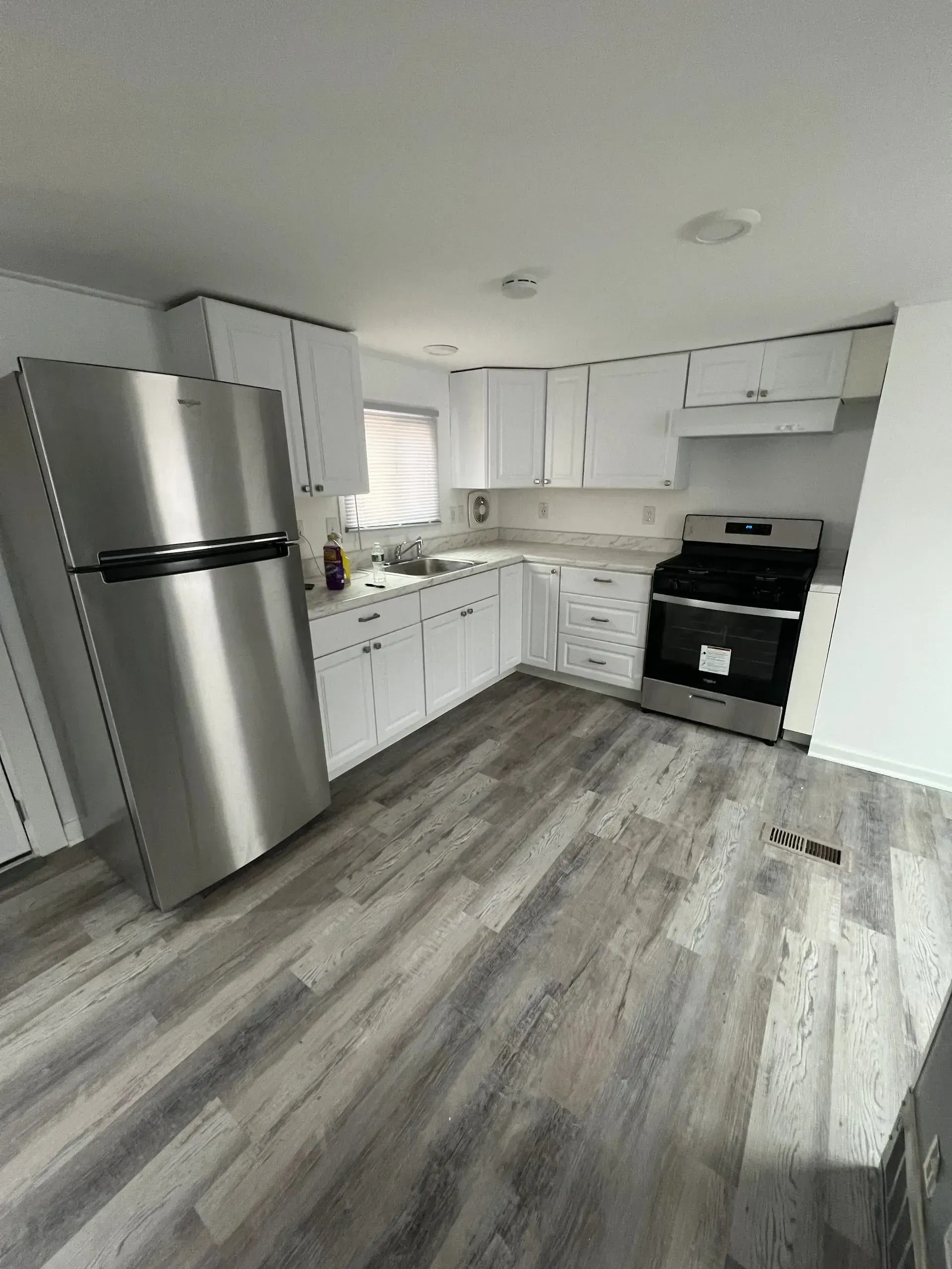Modern white kitchen with stainless steel refrigerator, black stove, and gray wood flooring.