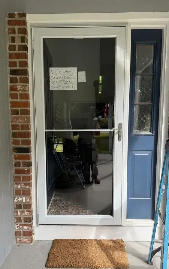 Front porch with a white screen door, narrow blue side panel, brick wall, and doormat.