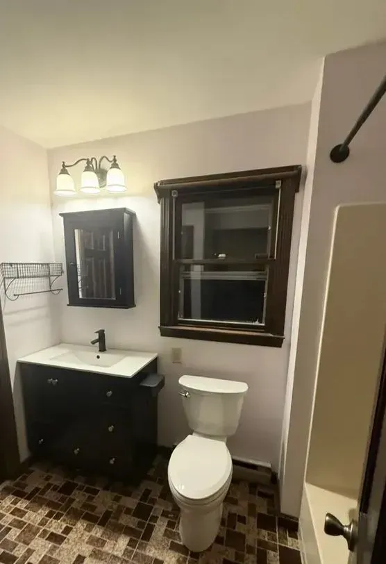 Small bathroom with dark vanity, mirror, window, and toilet under warm light.