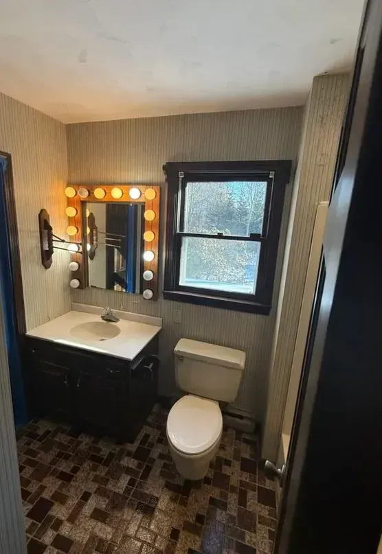 Small bathroom with vanity, lit mirror, toilet, and window, showing a narrow tiled space.