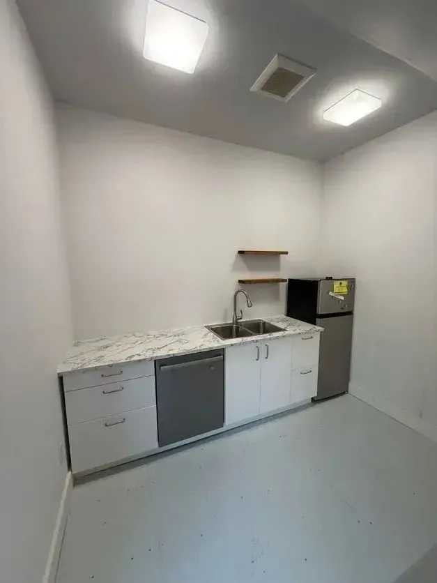 Small white kitchenette with sink, cabinets, refrigerator, and overhead lights in a plain room