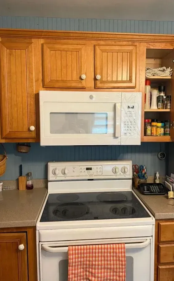 Kitchen with white microwave above a white electric stove, framed by honey-colored cabinets and blue wall.