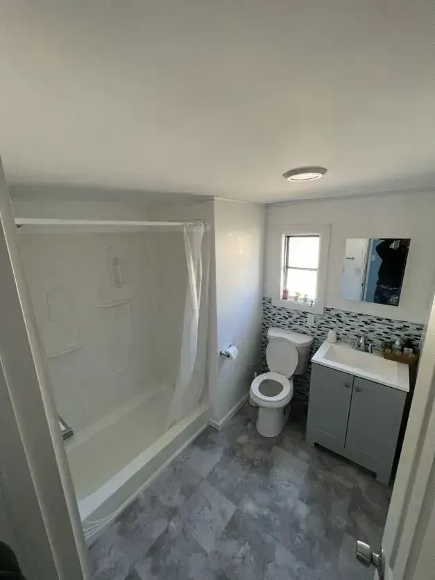 Small bathroom with shower-tub, toilet, vanity, window, and gray tile flooring.