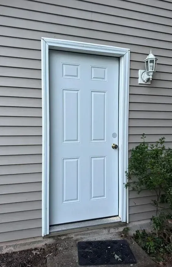 White front door with sidelights on a gray-sided house, with a porch light and small steps.
