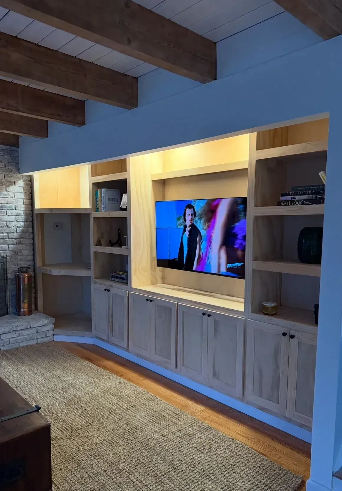 Built-in living room media wall with TV, illuminated shelves, and wood cabinets under exposed beams