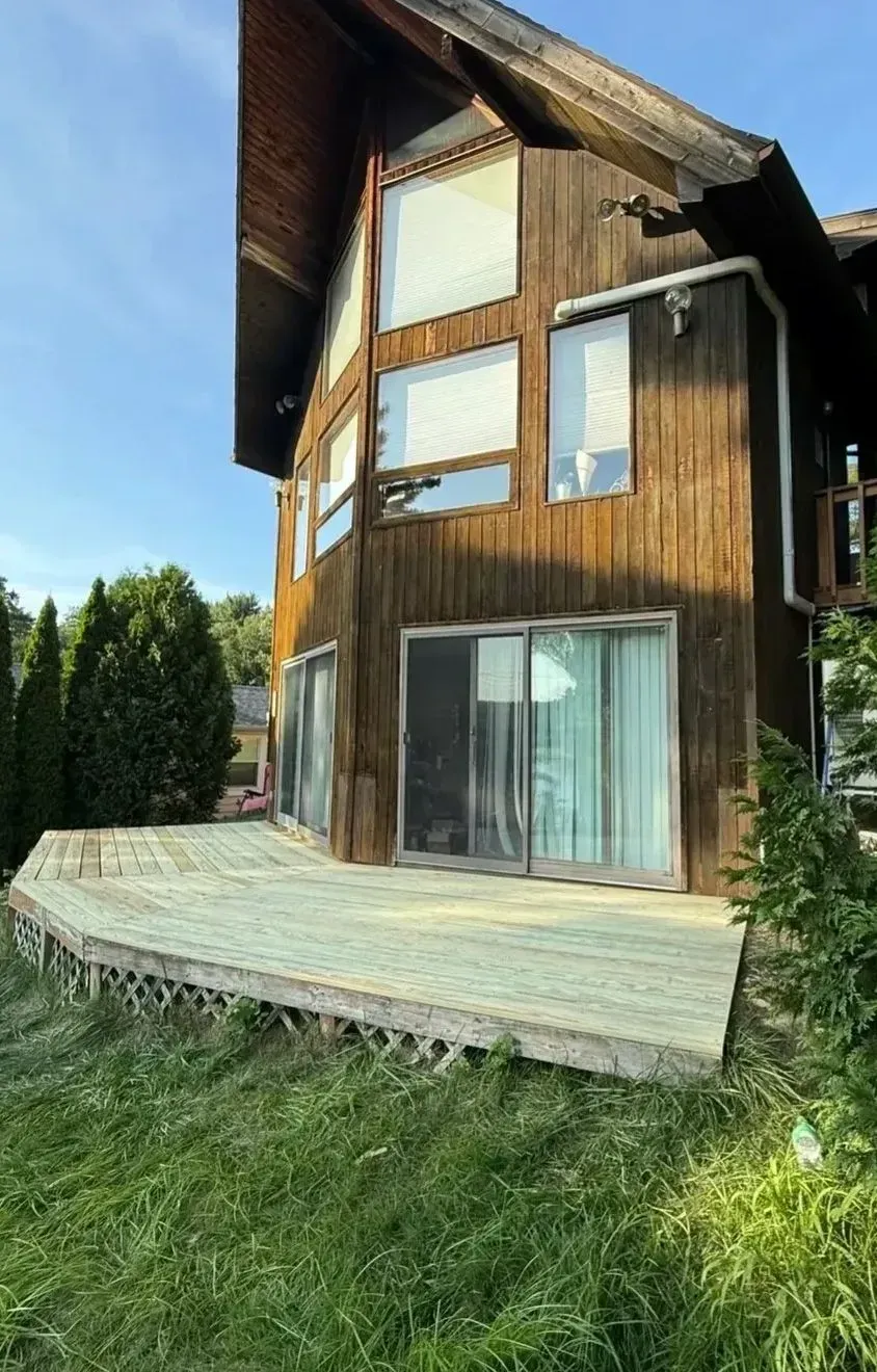 Wooden house with large windows and a curved deck, viewed from the lawn under a blue sky