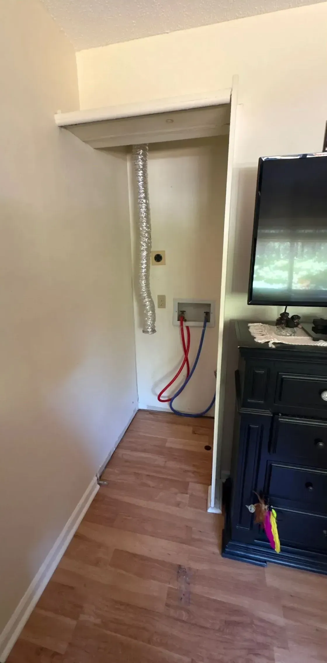 Narrow hallway with exposed cables, a wall opening, and a black cabinet in a wood-floored room.