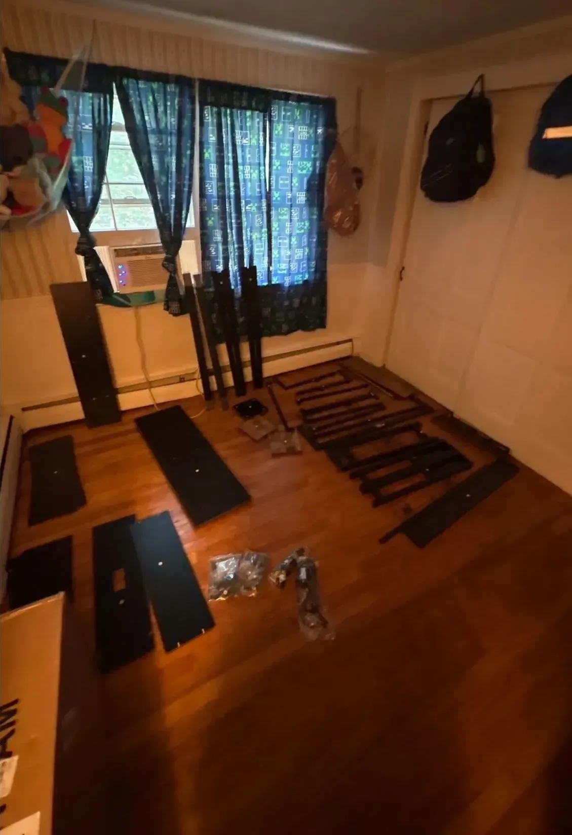 A small room with disassembled furniture parts laid on the wooden floor near a window with blue curtains.