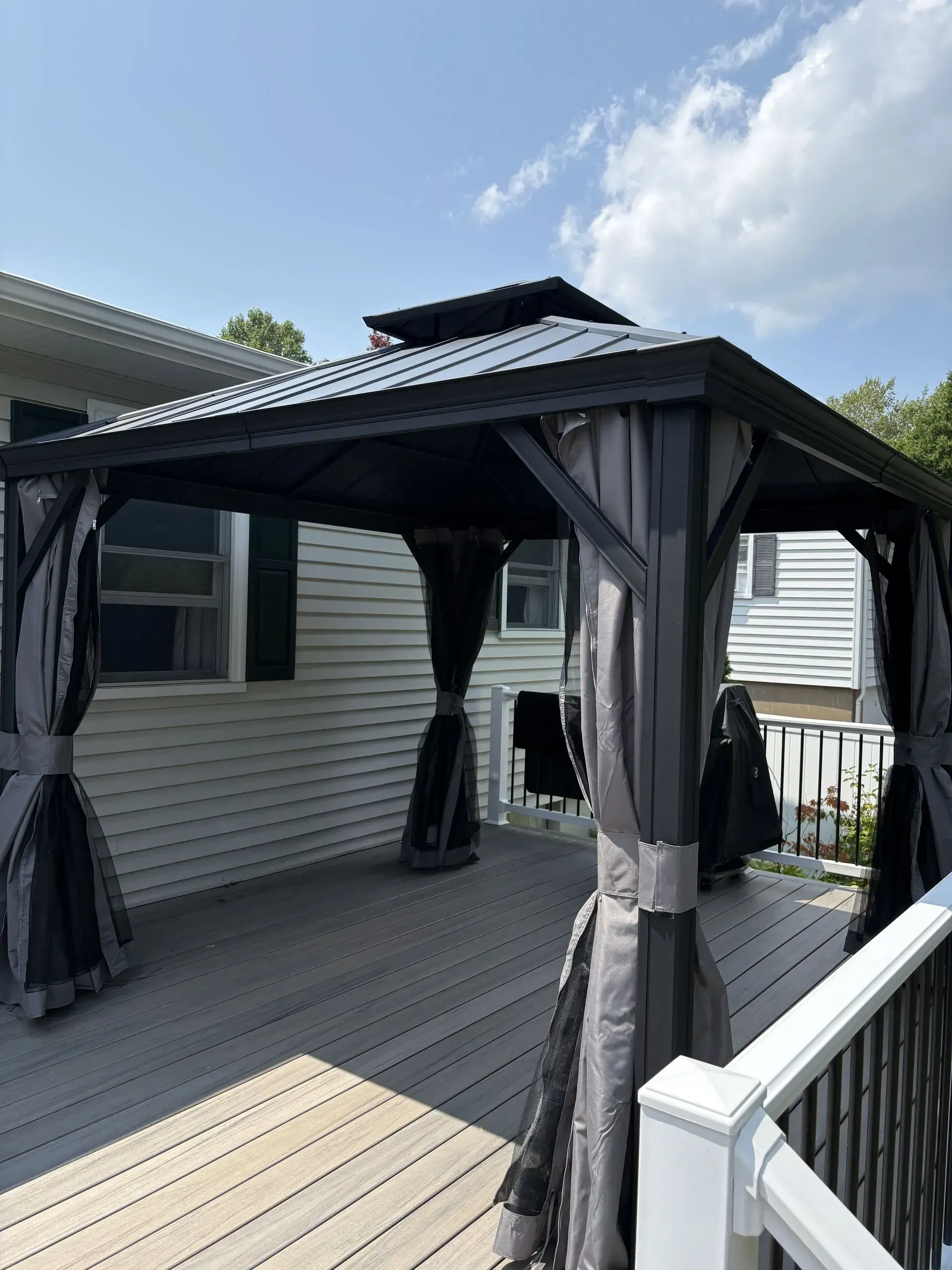 Covered backyard deck with a dark gray gazebo and railings beside a white house.