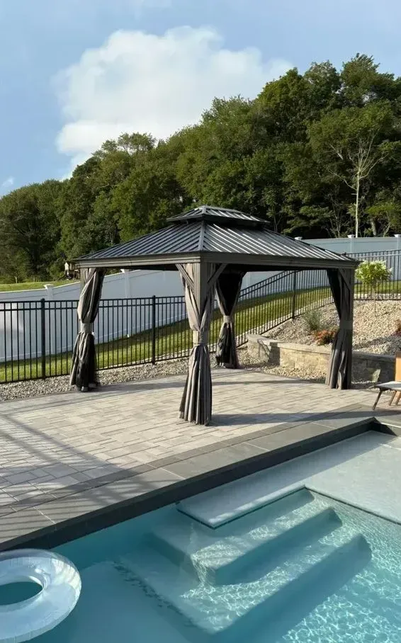 Backyard pool with a black gazebo, stone patio, and river view beyond the fence
