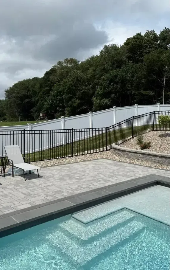 Patio with a small pool, lounge chair, and black railing beside a curving road and wooded hillside