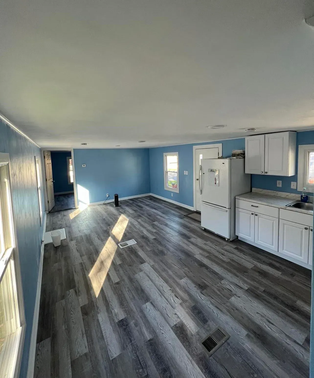 Empty open-concept room with gray floors, blue walls, white cabinets, and sunlight streaming in from the left.