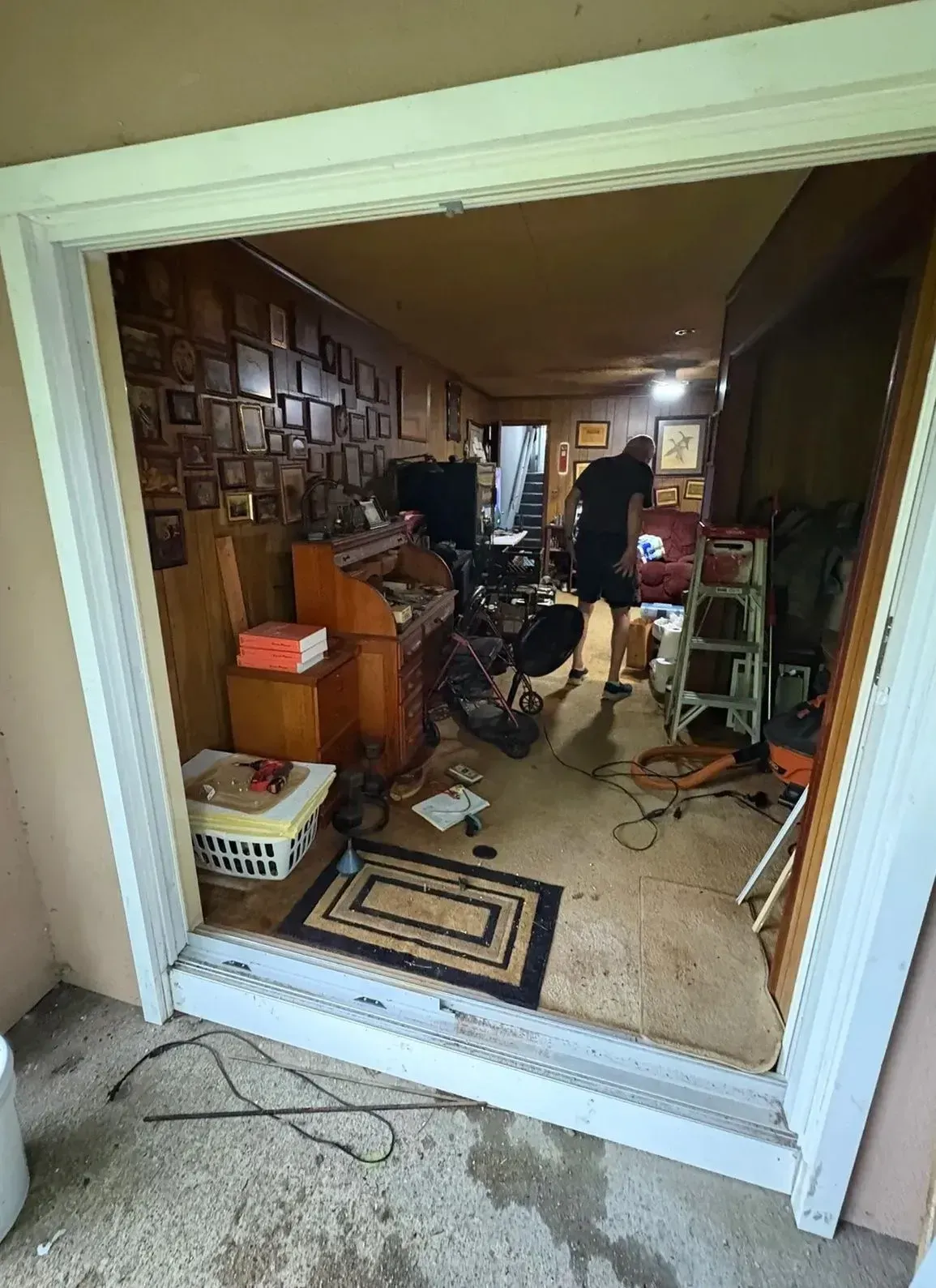 Cluttered room viewed through a doorway, with furniture, boxes, and a bicycle in a dim interior.