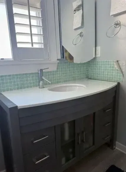 Modern bathroom vanity with white sink, dark cabinet, mint tile backsplash, and window above sink