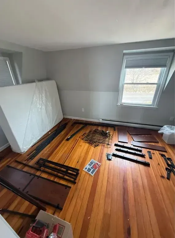 Room with hardwood floor, gray walls, a window, and stacked dismantled bed frame parts on the floor.