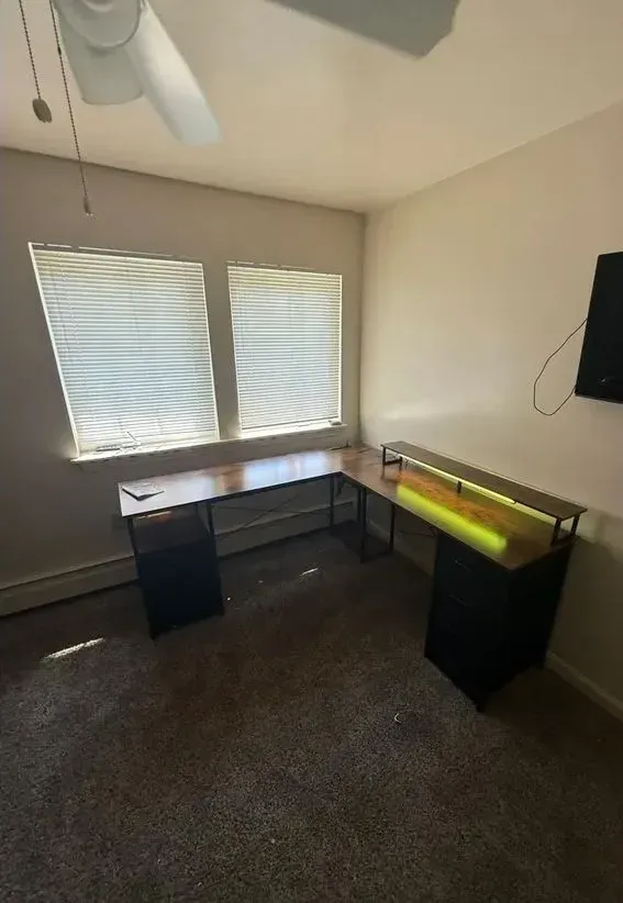 Empty home office with L-shaped desk, two windows with blinds, ceiling fan, and wall-mounted TV.