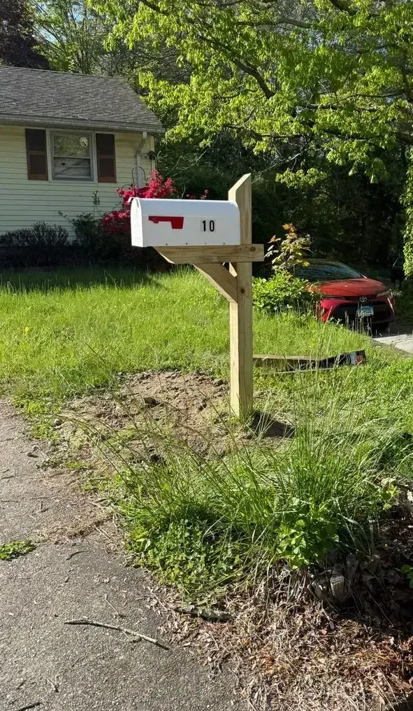 White mailbox on wooden post beside a grassy yard and driveway, with a house and red car in the background.