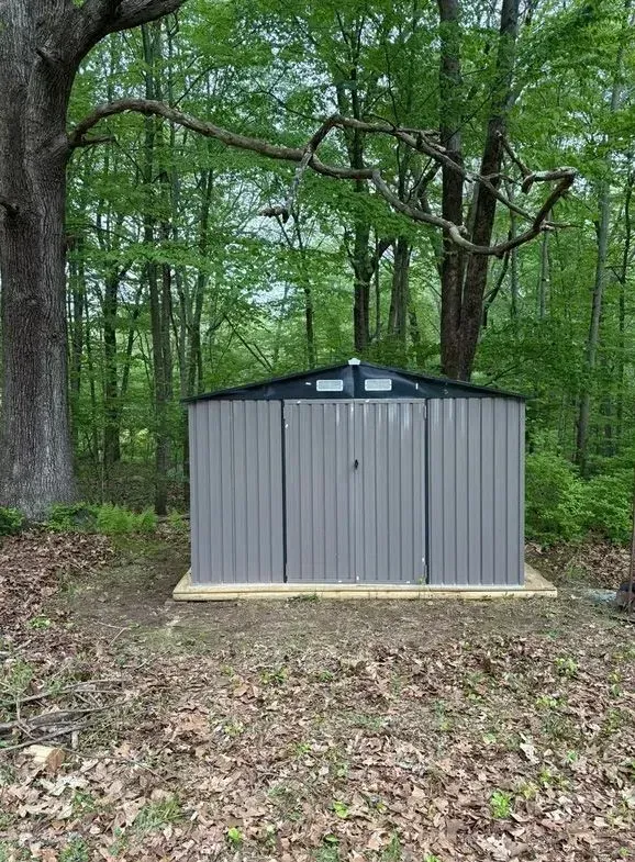 Gray metal shed in a wooded backyard, sitting on a concrete pad under tall trees.