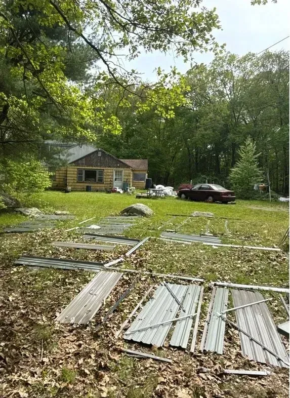Rural yard with a small cabin, red SUV, and corrugated metal sheets scattered among leaves and trees
